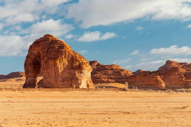 Dramatik bir çöl sahnesinde devasa bir Fil Kayası (Jabal Al Fil) kemeri ve katmanlı oluşumlar bulunur. Geniş düzlükler ve parlak bir gökyüzü yalnızlığı, keşfi ve doğal güzelliği çağrıştırıyor..