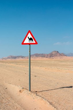 Geniş bir çöl ovasında, metal bir direğe tek başına geçen bir deve uyarı levhası yükselir. Kayalık dağlar ufukta parlayan mavi gökyüzü yalnızlık ve seyahat duygusunu güçlendirir..