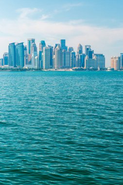 Ufuk boyunca uzanan modern Doha şehir merkezinin panoramik görüntüsü, önplandaki sakin mavi sulardan yansıyor. Parlak gündüz sahnesi şehir enerjisini, kıyı yaşamını ve mimari gelişmişliği taşır.