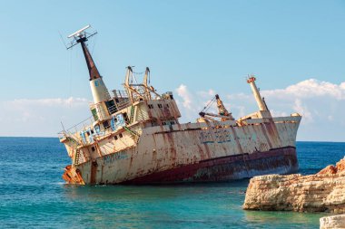 Paslı kargo gemisi Kıbrıs kıyılarında karaya vurmuş, pürüzlü kayalara yaslanmış. Parlak mavi gökyüzünün altında boya, pas ve durgun turkuaz sularla dramatik bir deniz sahnesi..