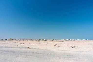 Katar, geniş, kurak bir çöl manzarası ufuk boyunca uzanan uzak bir kasaba silueti parlak, açık mavi bir gökyüzünün altında. Sessiz, geniş ve huzurlu, seyahat, manzara ve kentsel karşıtlık temaları için uygun.