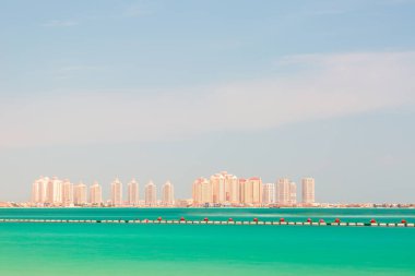 Doha, İnci Adası, Katar. Turkuaz su boyunca uzanan bir dizi modern yüksek binayla parlak bir sahil manzarası. Temiz bir gökyüzü ve sakin bir ufuk, seyahat ve lüks yaşam temaları için ideal bir deniz kenarı atmosferini çağrıştırıyor.