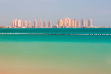 Doha, İnci Adası, Katar. Turkuaz su boyunca uzanan bir dizi modern yüksek binayla parlak bir sahil manzarası. Temiz bir gökyüzü ve sakin bir ufuk, seyahat ve lüks yaşam temaları için ideal bir deniz kenarı atmosferini çağrıştırıyor.
