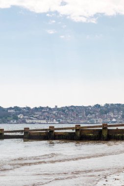 Swanage. Dorset, İngiltere. Sakin bir kıyı sahnesinde sakin bir denize uzanan yıpranmış bir dalgakıran uzak sahilde yoğun inşa edilmiş bir kasaba ve yukarıda soluk mavi bir gökyüzü..