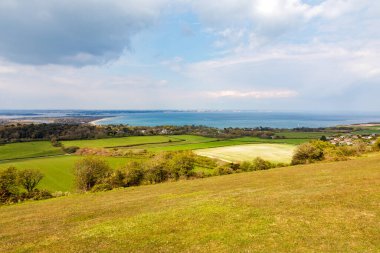 Studland köyü, Ballard Cliff yolu. Dorset. Zümrüt tarlaları, dağınık ağaçlar, uzak bir köy ve ufuk çizgisinde bulutlu bir gökyüzünün altında sakin bir mavi deniz içeren sakin bir kıyı manzarası. Seyahat, doğa ve yaşam tarzı için mükemmel.