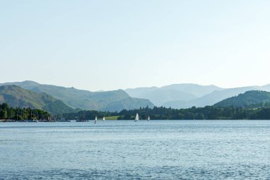 Ullswater, Göl Bölgesi. Yuvarlanan yeşil tepeler ve uzak dağlarla çerçevelenmiş sakin bir göl manzarası. Küçük yelkenliler sakin sularda süzülür parlak gökyüzünün altında, huzurlu bir açık hava eğlencesi, seyahat ve doğal manzara güzelliği uyandırır..