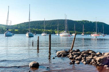 Ullswater, Göl Bölgesi. Arka planda yeşil tepeler ve sakin suyla kayalık bir sahile demir atmış birkaç yelkenlinin yer aldığı sakin bir liman sahnesi. Denizcilik, seyahat ya da açık hava yaşam tarzı görüntüleri için mükemmel..