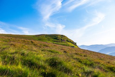 Ullswater, Göl Bölgesi. Güneşli bir yamaçtaki çayır, açık mavi gökyüzünün altındaki uzak bir sırta doğru uzanır. Yuvarlanan yeşiller ve sakin manzaralar açık hava keşiflerini ve barışçıl, açık arazi kaçışlarını davet ediyor..