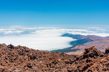 Teide Dağı manzarası - Önünde engebeli kayalar, altında bulut denizi ve yukarıda açık mavi bir gökyüzü olan engin volkanik zirve manzarası. Seyahat, macera, doğa ve manzara fotoğrafçılığı için ideal..