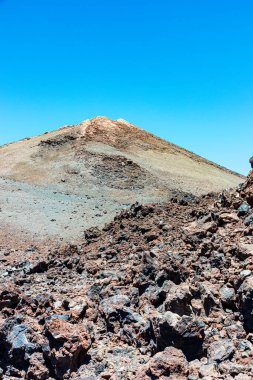 Katıksız bir volkanik Tenerife manzarası, canlı mavi bir gökyüzünün altında, eğimli Teide Dağı zirvesi, kumlu kayalık ön plan ve çok renkli mineral katmanlarını barındırır. Keşif, macera, doğa, izolasyon ve aşırı arazi temaları için ideal.
