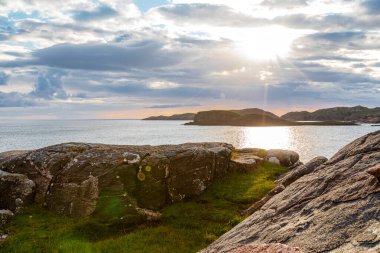 İskoçya İskoçya - Oldhoremore - engebeli kayalar, yeşil çimenler ve sakin bir deniz içeren sakin bir kıyı sahnesi. Güneş ışınları bulutların arasından çıkar, uzak adaları aydınlatır ve sakin bir doğa anı için yansıtıcı bir su yüzeyi oluşturur.