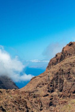 Parlak mavi bir gökyüzünün altında engebeli kaya yüzeyleriyle dramatik bir kıyı kayalığı ön planda hüküm sürüyor. Uzakta, okyanus ufka doğru uzanıyor, sürüklenen bulutlar ve güneşli ışıkla çerçevelenmiş..