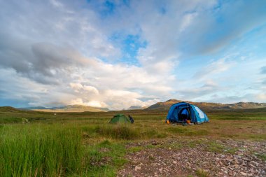 İskoçya Highlands NC500 - sakin bir açık hava kamp sahnesi uzak dağlarla geniş bir çim ovasına kurulmuş mavi ve yeşil çadırları gösteriyor. Parlak bulutlar ve mavi gökyüzü dramatik bir zemin oluşturur, davetkar macera, keşif.