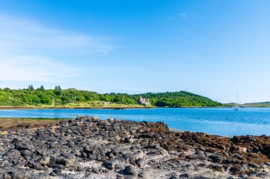 Dunvegan Gölü - Sakin bir sahil sahnesi kayalık bir ön plan, şato harabeleri ile bereketli bir orman adası, ve berrak bir gökyüzü altında sakin mavi su üzerinde uzak bir yelkenli..