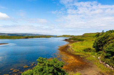 Loch Dunvegan - Yeşil tepeleri, ağaçları ve parlak mavi gökyüzünün altındaki kayalık kıyı şeridini gösteren sakin bir göl sahnesi. NC500 macerası. Huzurlu, doğal manzara doğa, seyahat ve açık hava görüntüleri için mükemmel..