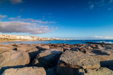 Kosta Adeje kenti ufuk boyunca yükselirken, altın güneş ışığı kayalık bir kıyı şeridini aydınlatıyor. Sakin bir deniz ve mavi gökyüzü seyahat, tatil ve şehir sahili temaları için ideal bir deniz manzarası yaratır..