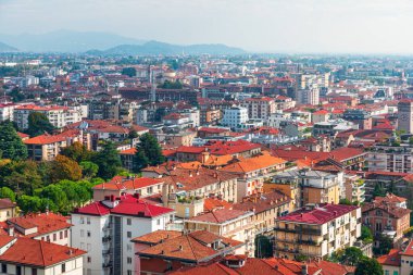 Kırmızı kiremitli ve çeşitli orta katlı binalarla dolu hareketli bir Bergamo mahallesinin geniş bir hava manzarası. Ağaçlar yemyeşil caddeleri doldururken uzak tepeler ufuk çizgisini çerçeveletip şehir yaşamını, sıcaklığı ve güneş ışığını yansıtıyor..