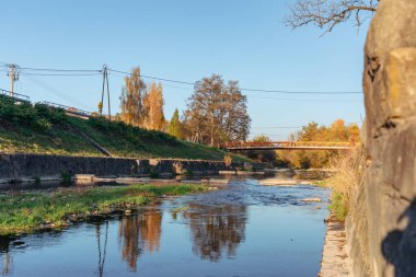 Huzurlu bir nehir manzarasında Grybow 'daki Biala Nehri' nin sığ bir kanalı, uzak bir yaya köprüsü, taş setler ve suya yansıyan sonbahar ağaçları yer alır. Sıcak güneş ışığı kırsal manzarayı ve şehir yaşamına olan bağlantıyı vurguluyor.