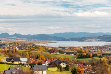 Sakin bir sonbahar manzarasında sakin bir göl kenarındaki dalgalı tarlalara dağılmış küçük bir köy, renkli ağaçlar, uzak dağlar ve parlak, geniş bir gökyüzü, barışı ve kırsal cazibeyi çağrıştırıyor..