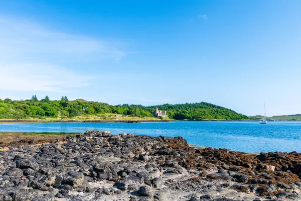 Dunvegan Gölü - Sakin bir sahil sahnesi kayalık bir ön plan, şato harabeleri ile bereketli bir orman adası, ve berrak bir gökyüzü altında sakin mavi su üzerinde uzak bir yelkenli..