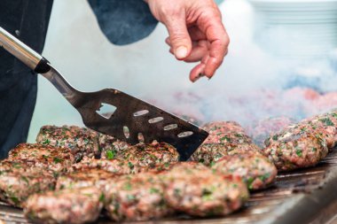 Sulu, baharatlı sığır burgerleri dumanlı ızgarada cızırdarken bir el spatulayla köfteleri çeviriyor. Güneşli bir açık hava piknik ortamında Aromalar yükselir, lezzetli, toplu ızgara anını yakalar..