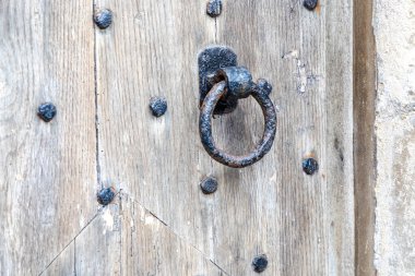 Yıpranmış ahşap bir kapıya yapıştırılmış eski, paslanmış demir kapı tokmağı. Dairesel halka ve perçinlenmiş çiviler tarihi cazibeyi ve kırsal, zamansız dokuyu çağrıştırıyor..