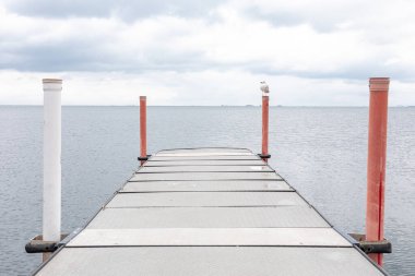 Kırmızı ve beyaz direklerle çevrili sakin suya uzanan metal bir rıhtım var. Yalnız bir martı, gri bulutlar üzerinde sürüklenirken bir direkte oturur ve huzur dolu, derin düşüncelere dalmış bir sahne yaratır..