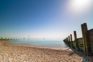 Bir çakıl taşı plajı ve durgun mavi sulara uzanan yıpranmış ahşap bir dalgakıranın yer aldığı sakin bir sahil sahnesi. Selsey Doğu Sahili. Doğa, seyahat ve deniz kenarı huzur temaları için ideal.