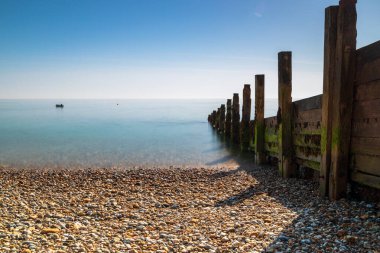 Bir çakıl taşı plajı ve durgun mavi sulara uzanan yıpranmış ahşap bir dalgakıranın yer aldığı sakin bir sahil sahnesi. Selsey Doğu Sahili. Doğa, seyahat ve deniz kenarı huzur temaları için ideal.