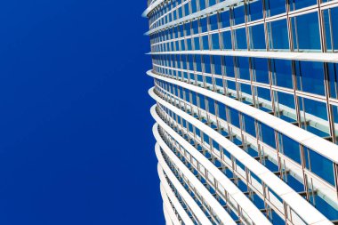 Londra, İngiltere: 28 Şubat 2025. Pürüzsüz, modern bir gökdelen, parlak mavi gökyüzüne karşı beyaz eğimli bantlar ve yansıtıcı mavi pencereler içerir. Bu mimari detay b için çağdaş tasarımı, simetriyi ve kentsel karmaşıklığı vurgular.