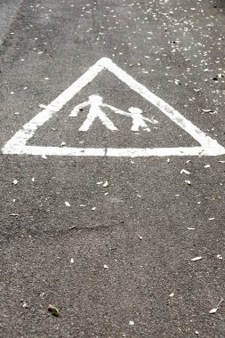 Siyah asfalt üzerinde beyaz üçgen bir yaya geçidi. İçinde yetişkin ve çocuk figürleri var. Olay yeri, güvenlik, ihtiyat ve dağınık yapraklı bir okul veya şehir bağlamını iletir..
