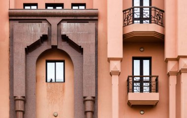 Kalın kemerler, geometrik detaylar ve metal balkon korkulukları içeren şeftali rengi bir bina cephesi. Yalnız bir kuş pencereden seyrediyor, şehir mimarisine sessiz ve düşünceli bir hava katıyor..