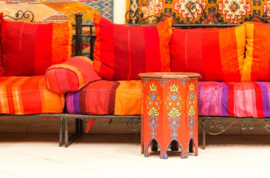 Kırmızı, turuncu ve mor yastıklı işlenmiş demirden bir kanepenin yer aldığı Fas 'ın canlı oturma alanı. Süslü turuncu bir masa ve mavi çiçekli, sıcak, cezbedici bir Ortadoğu atmosferi yaratıyor..