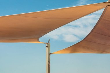 Güneş ışığı yelkenleri, açık hava direğinin üzerinde modern bir tepe örtüsü oluşturur. Parlak mavi gökyüzü ve kıvrımlı kumaş aksanlarıyla sakin ve havadar bir manzara yaratır..