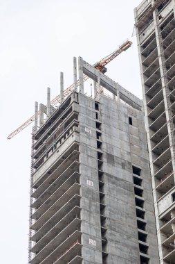 Yoğun bir inşaat sahasında, yukarıdaki vinçlerle desteklenen yüksek bir beton iskelet yükseliyor. Görüntü kentsel gelişimi, sanayiyi ve modern mimarinin ilerlemesini aktarır..