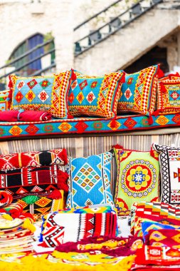 Canlı, el yapımı kilim yastıklar ve koyu maviler, kırmızılar ve sarılar sergileniyor. Dış pazar ortamında gösterilen sahne, zengin geometrik desenleri ve geleneksel tekstil işçiliğini vurguluyor.