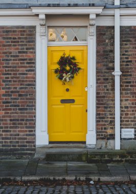 Beyaz süsleme ile çerçevelenmiş, neşeli bir Noel çelengiyle süslenmiş canlı sarı bir ön kapı. Tuğla cephesi ve kaldırım taşı basamakları hoş bir bayram cazibesini ve eve hoş geldin hediyesini çağrıştırıyor..