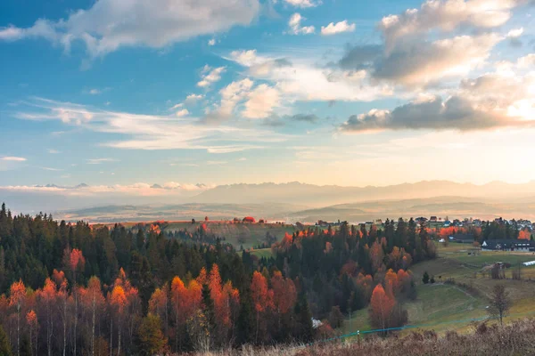 Parlak bir sonbahar Nowy Targ arazisi yama işi bir orman ve uzak Buflak köyü ve Tatras dağları ile yuvarlanan tepeler üzerinde. Ilık altın saat ışığı, açık mavi gökyüzü altında kırmızıları, portakalları ve yeşillikleri artırarak sakin bir kırsal ortam yaratıyor..