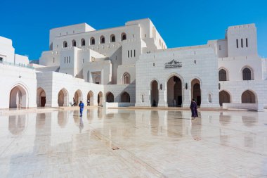Royal Opera House Muscat (ROHM), Muscat, Oman: December 31, 2023. A grand white architectural complex of the Royal Opera House Muscat featuring arches and a expansive courtyard, reflecting in the polished marble surface. Visitors walk the sunlit plaz