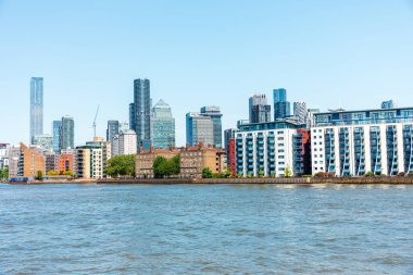 Londra, İngiltere: 11 Mayıs 2025. Modern bir Canary Wharf 'ın panoramik manzarası, Londra' nın nehir kıyısı boyunca uzanan ufuk çizgisi, cam gökdelenler, deniz manzaralı binalar ve yemyeşil alanlar. Parlak mavi gökyüzü kentsel enerji, büyüme ve şehir iletir