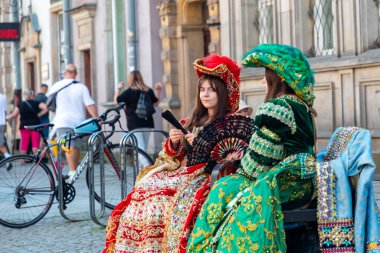 Gdansk, Polonya: 2 Haziran 2024. Canlı, süslü elbiseler ve dekoratif başlıklar giyen iki genç kadın kalabalık bir şehir caddesinde bisikletlerin yanındaki bankta oturuyor. Sahne, tarihten esinlenilmiş moda ile şehir yaşamı ve sokak fotoğrafçılığı enerjisini harmanlıyor..