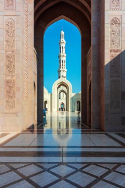 Sultan Qaboos Ulu Camii, Muscat, Umman - 31 Aralık 2023: Karmaşık oyulmuş taş duvarlar tarafından çerçevelenmiş muazzam bir cami kemeri ve minare manzarası. Sıcak kumtaşı tonları, zarif simetriyi ve zamansız yayı yansıtan açık mavi gökyüzü ile buluşur.