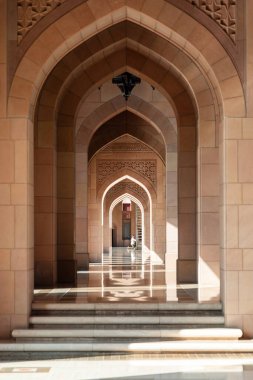 Sultan Qaboos Büyük Camii, Muscat, Umman - 31 Aralık 2023: Sıcak taş kemerlerden oluşan uzun, güneşli bir koridor ritmik bir perspektif yaratır. Cilalı zemin kemerleri yansıtıyor, sükuneti, zarafeti ve mimari zarafeti arttırıyor..