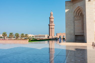 Sultan Qaboos Büyük Camii, Muscat, Umman - 31 Aralık 2023: Uzun bir minare, kemerli kapı aralığı, geniş mermer plaza, palmiye çizgili bahçeler ve açık mavi gökyüzünün yer aldığı dingin bir İslami mimari sahne. Ziyaretçiler yansıtıcı yüzeyde yürür.