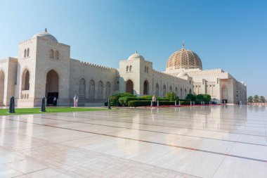Sultan Qaboos Büyük Camii, Muscat, Umman - 31 Aralık 2023: Kubbeli çatıları, soluk taş duvarları ve zarif kemerleri olan büyük bir İslami cami geniş bir mermer plazanın yanında yer alır. Palmiye ağaçları ve manikürlü yeşillik, zamansız yaşam için sakin bir zemin sağlar.