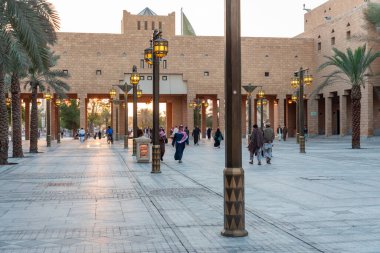 Riyad, Suudi Arabistan - 22 Aralık 2025: Sunlit Plaza 'da insanlar gün batımında geniş bir avluda yürürken palmiye ağaçları ve taş kemerleri halka açık bir meydanda sakin ve kültürel bir ortam yaratıyor..
