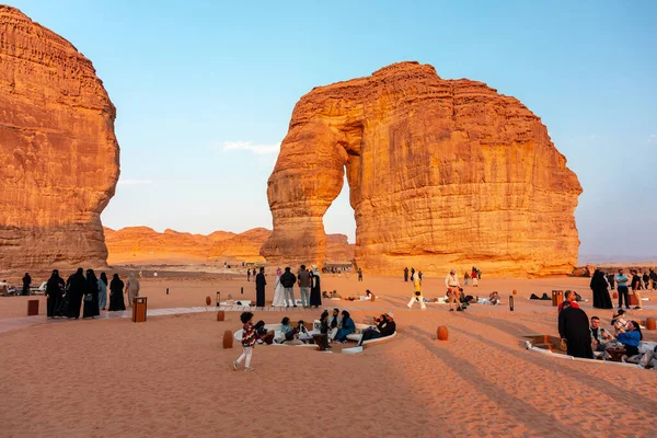 Fil Kayası, Alula, Suudi Arabistan - 26 Aralık 2025: Ziyaretçilerin toplandığı, dinlendiği ve sıcak altın saatin keyfini çıkardıkları devasa Fil Kayası kemerinin etrafında çöl gün batımı. Seyahat, kültür ve manzaralı çöl atmosferiyle dolu sosyal, açık bir sahne..