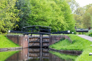 Bansigstoke Kanalı 'ndaki su kapıları. Surrey 'deki St. John köyü yakınlarındaki Goldsworth Park' taki Lush Green Trees ve Still Water Reflectionat yürüyüş yollarıyla çevrili tarihi kanal kilidi kapıları ve Footbridge. Bahar zamanı, yeşil ağaçlarla güneşli bir günde.