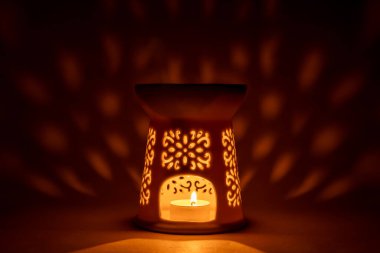 Beyaz seramik mum lambası ve içinde yanan bir çay ışığı. Arka planda güzel yumuşak ışık yansımaları, rahat ve atmosferik iç fotoğraf.