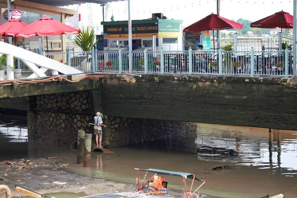 A local man fishing under a small bridge along the riverside near a waterfront cafe and ticket office in Hochiminh City, Vietnam. A realistic urban lifestyle scene showing contrast between leisure business above and traditional fishing activity below